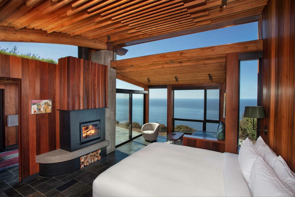 Post Ranch Inn bedroom interior with red wood walls, a cosy fireplace, and ocean views.